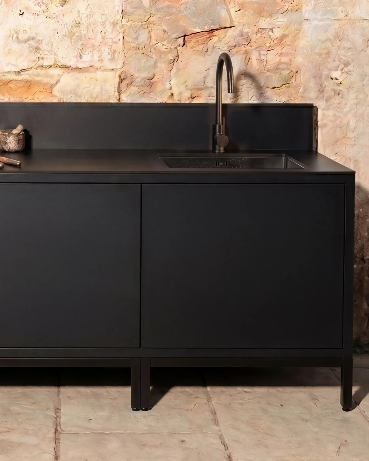 Módulo de esquina negro PILOTIS con fregadero y grifo, cocina exterior junto a pared de piedra