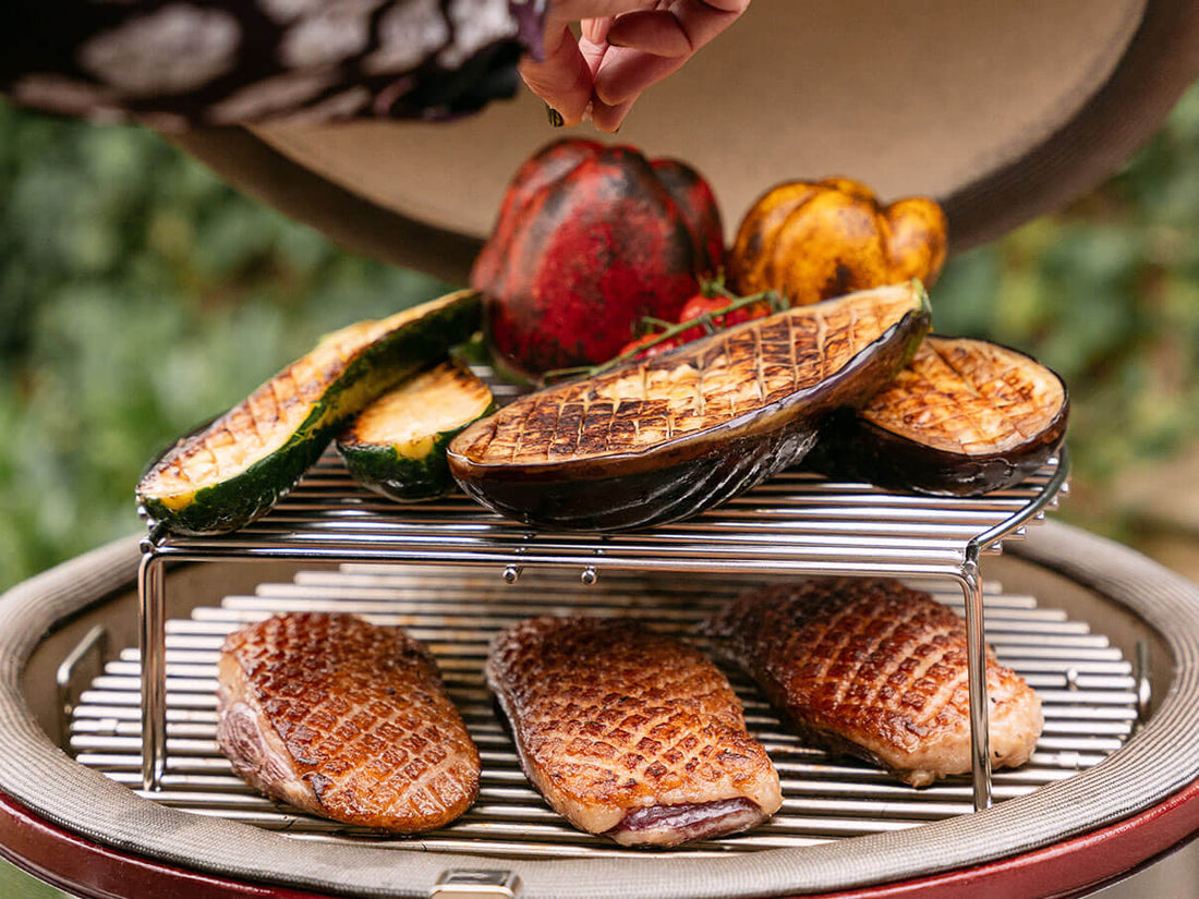 Cocción a dos niveles en un kamado Kokko con magrets de pato marcados en la parrilla inferior y verduras asadas en la parrilla elevada, ilustrando la versatilidad de cocción.