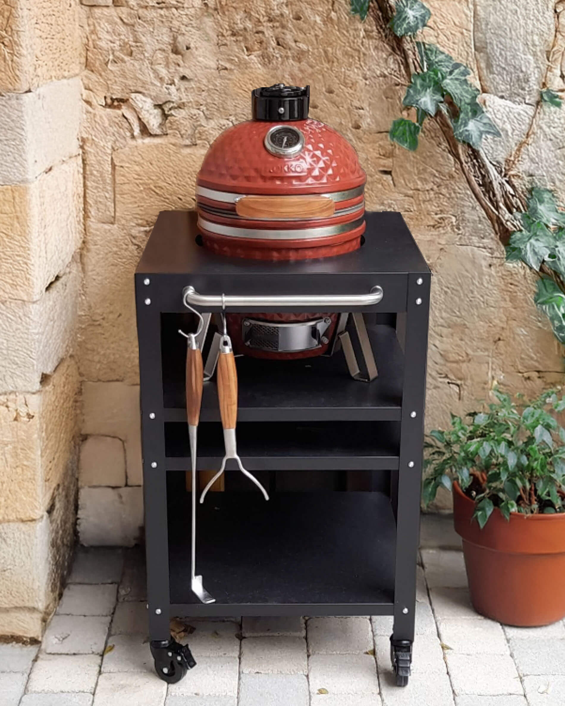 Kamado Kokko rojo de cerámica colocado sobre un carro negro con ruedas, con estante superior, espacio de almacenamiento abierto y utensilios colgados en la parte frontal, presentado en un patio de piedra.