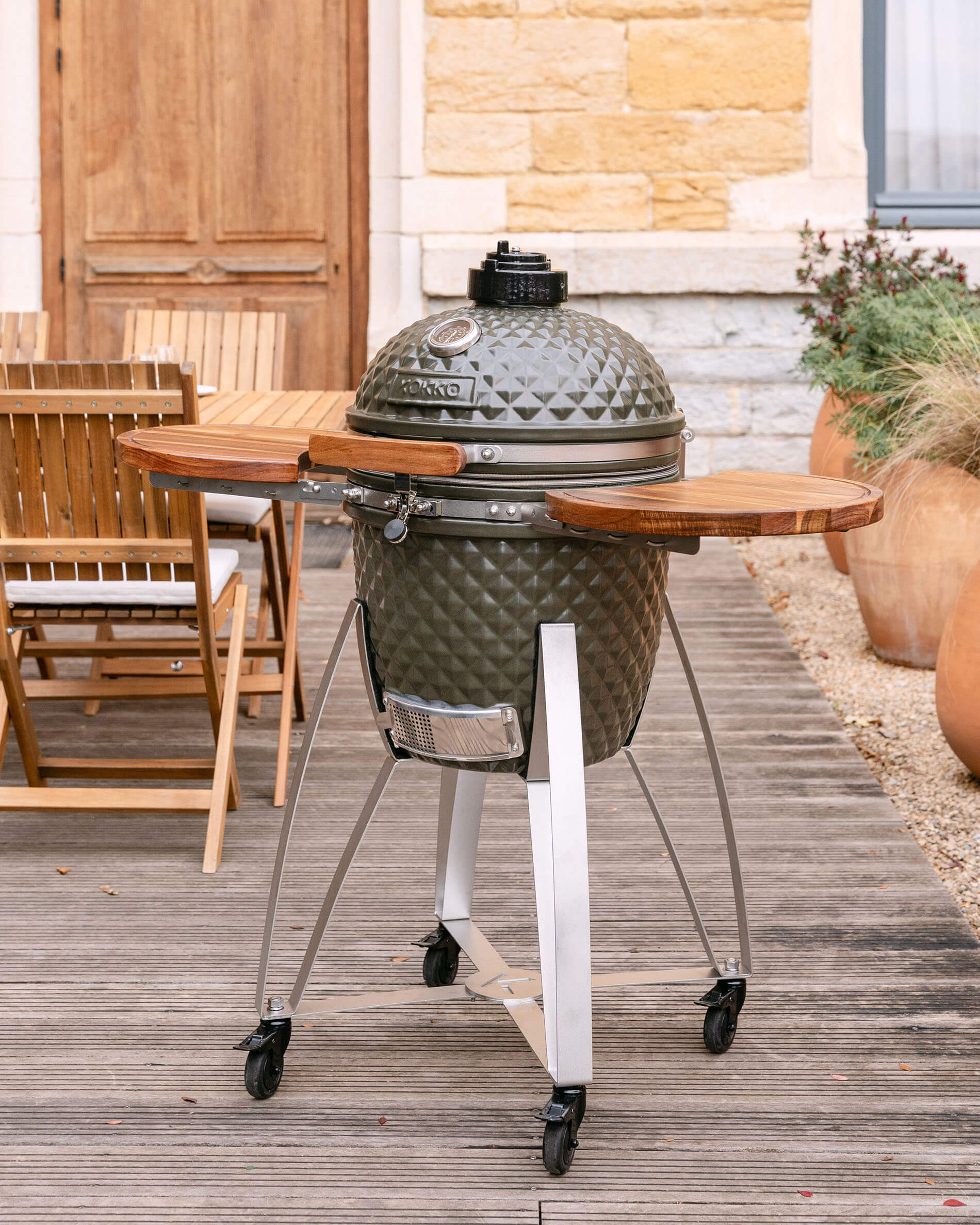 Kamado Kokko sobre carro móvil con estantes laterales de madera, presentado en uso exterior en una terraza.