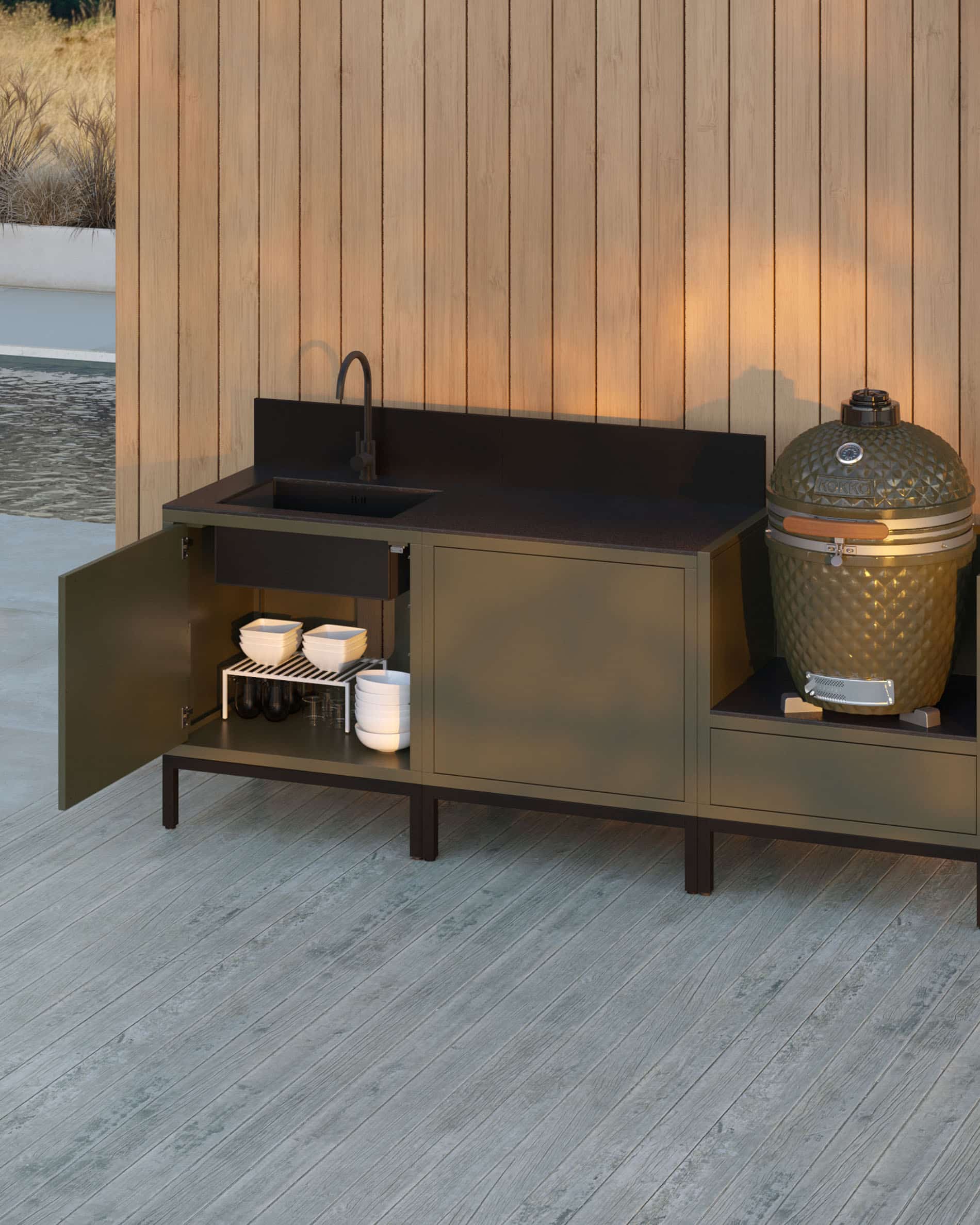 Cocina exterior pilotis de 3 módulos para kamado con fregadero integrado, mueble en color oliva con encimera y panel trasero negros, puerta del módulo izquierdo abierta mostrando un espacio de almacenamiento interior, fregadero con grifo a la izquierda, dos módulos centrales cerrados y kamado integrado en el módulo lateral derecho, estructura sobre patas negras, instalación en exterior junto a una pared revestida de madera.