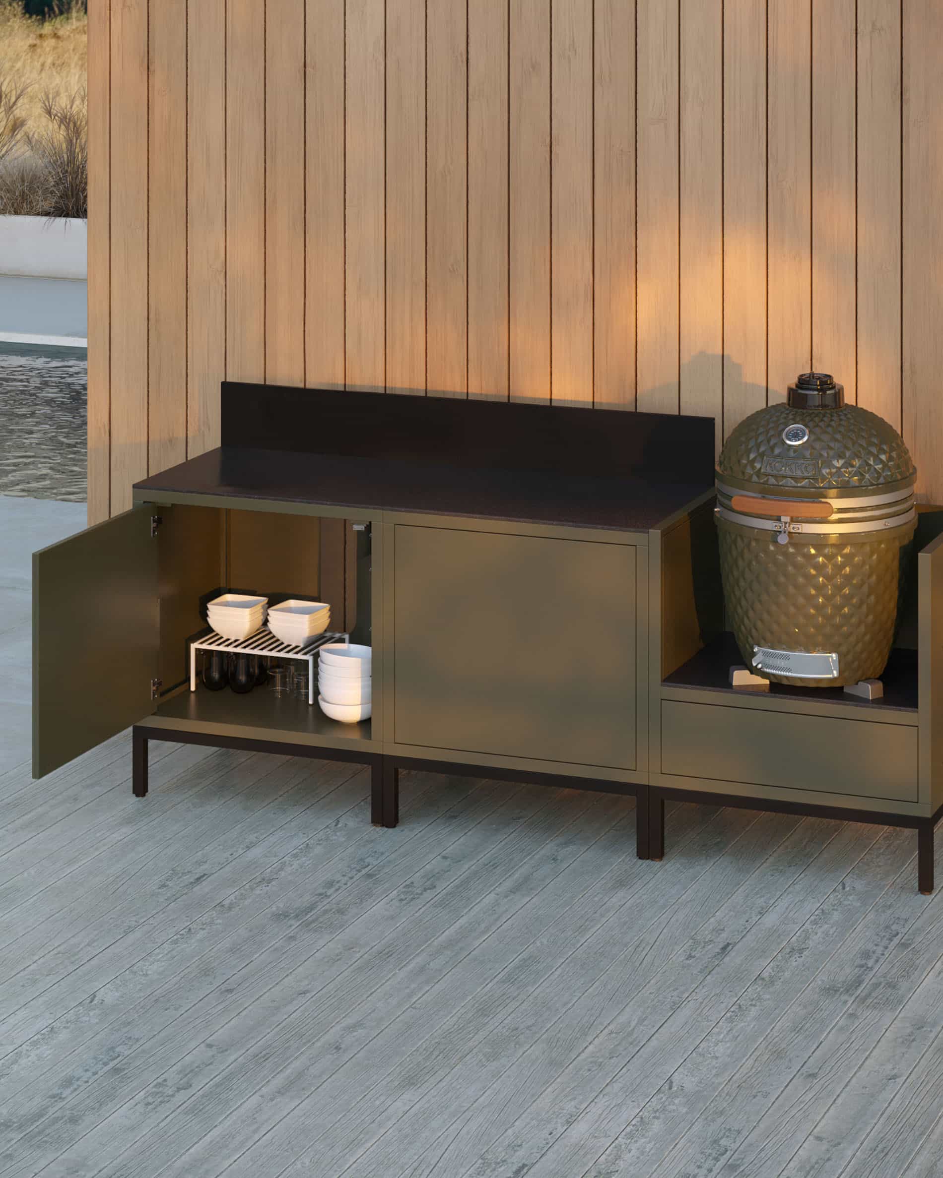 Cocina exterior pilotis de 3 módulos para kamado, mueble en color verde oliva con encimera oscura y panel trasero a juego, puerta de almacenamiento abierta mostrando vajilla guardada, kamado instalado en el módulo lateral derecho, estructura sobre patas negras.