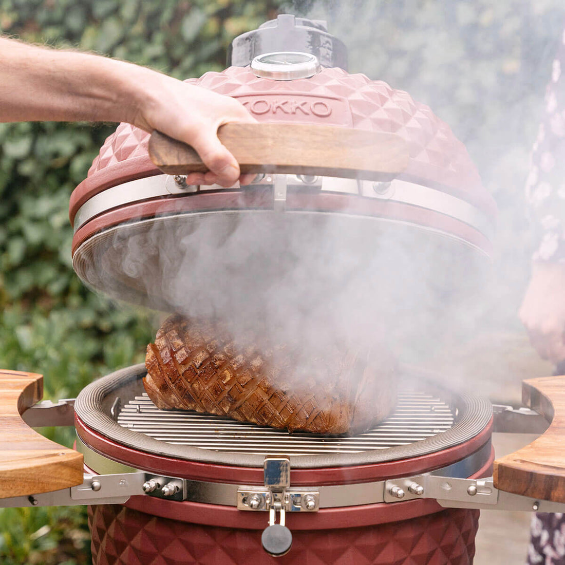 Una mano levanta la tapa de un kamado Kokko rojo óxido, dejando ver un trozo de carne asada sobre la parrilla de acero inoxidable. Una densa humareda sale de la cuba, resaltando la cocción al fuego de leña, la junta perimetral y las bandejas laterales de madera.