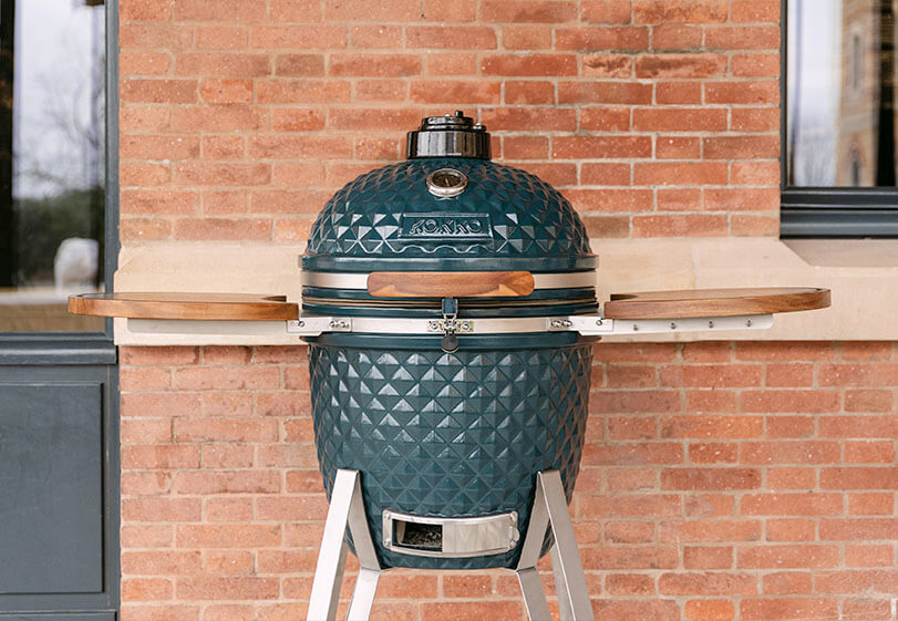 Kamado Kokko azul océano colocado sobre soporte con estantes de madera, presentado de frente en exterior junto a una pared de ladrillo.