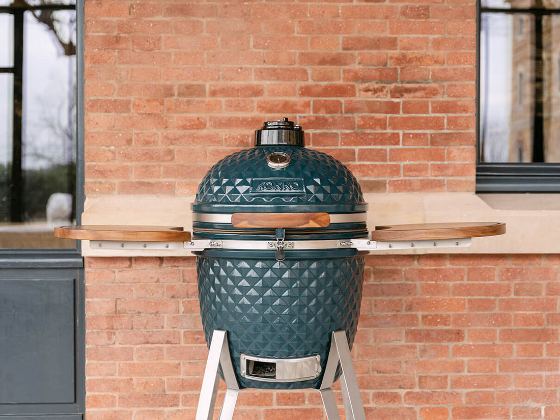 Kamado Kokko color verde océano, instalado en exterior frente a una pared de ladrillo. El kamado está cerrado, con una tapa de cerámica texturizada, un termómetro integrado en la parte superior, un asa de madera, un cierre de seguridad de acero inoxidable y dos bandejas laterales de madera de acacia. Todo el conjunto descansa sobre un soporte metálico con bandeja de ventilación inferior visible.