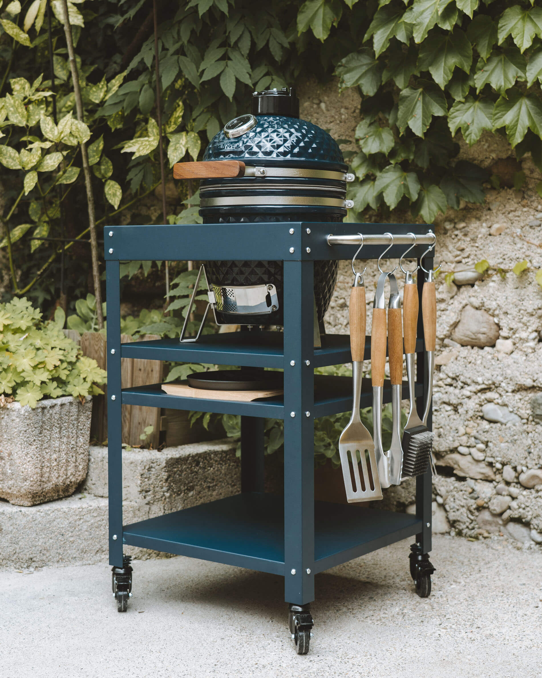 Kamado Kokko en color azul océano colocado sobre un carro metálico a juego, con estantes de almacenamiento, recogedor de cenizas y utensilios colgados en el lateral.