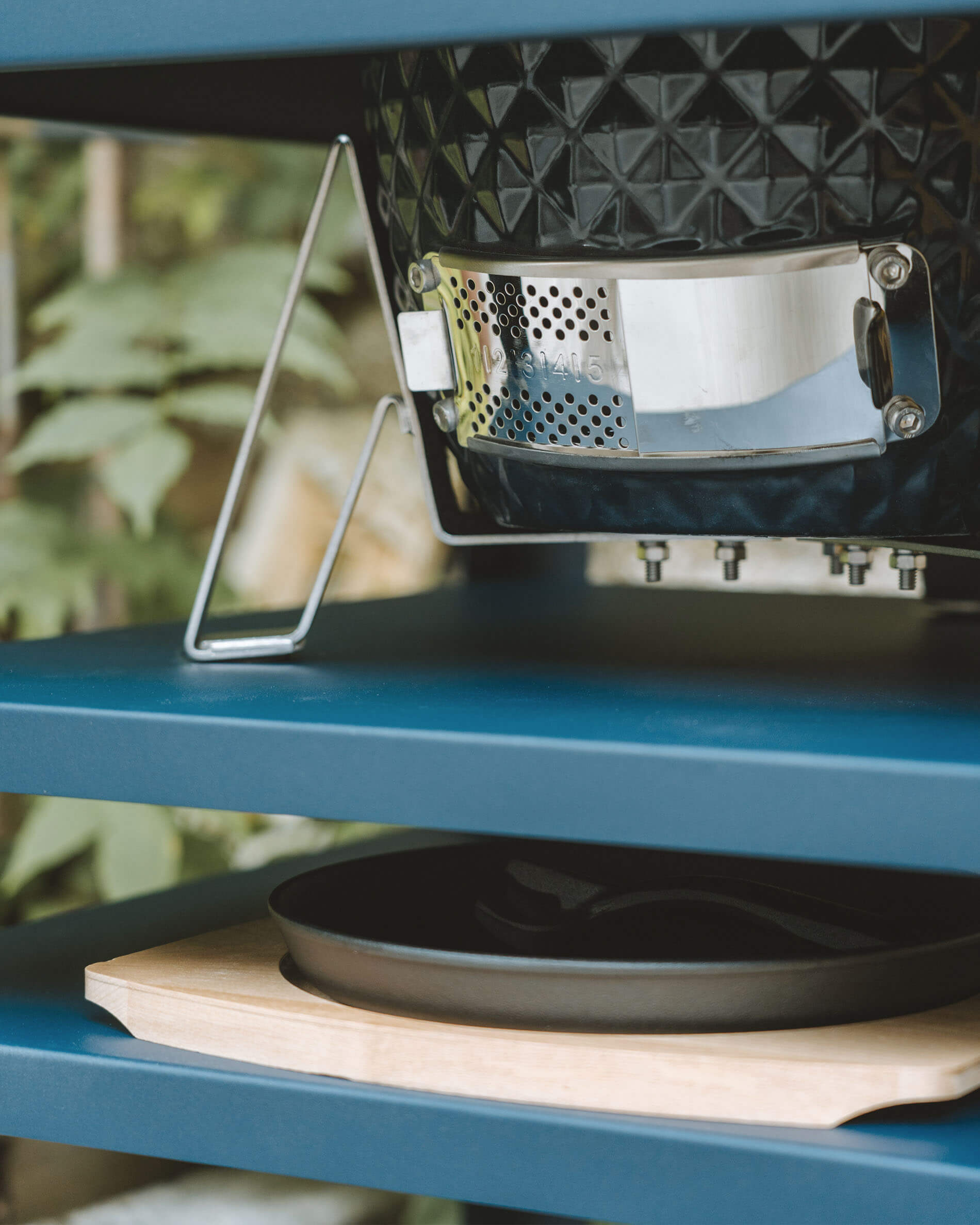 Primer plano de la ventilación inferior de acero inoxidable de un kamado Kokko, integrada en una mesa auxiliar metálica con bandeja recogecenizas.