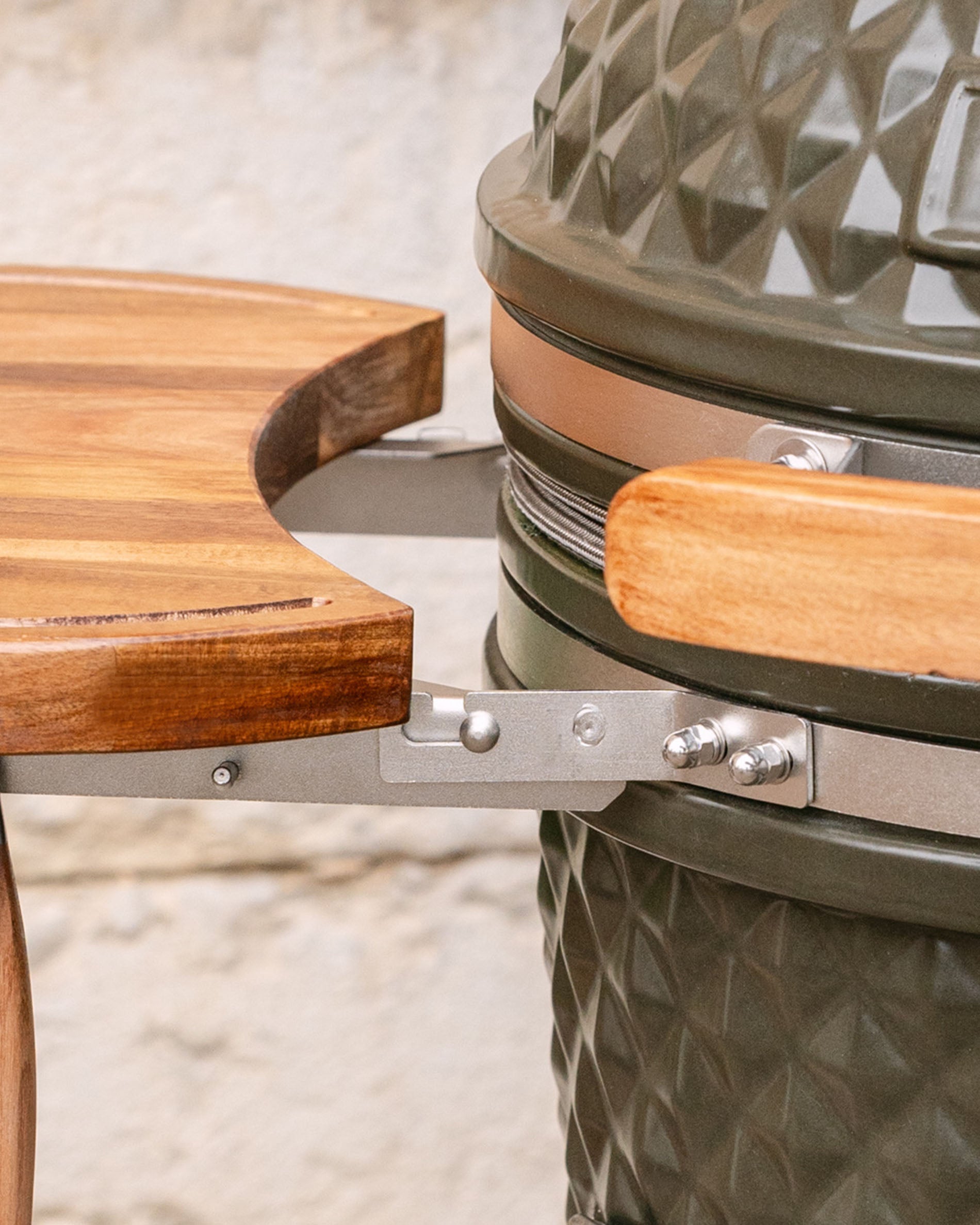 Detalle de las fijaciones metálicas que unen la bandeja lateral de madera al kamado, con asa de madera y aro de acero inoxidable visibles.