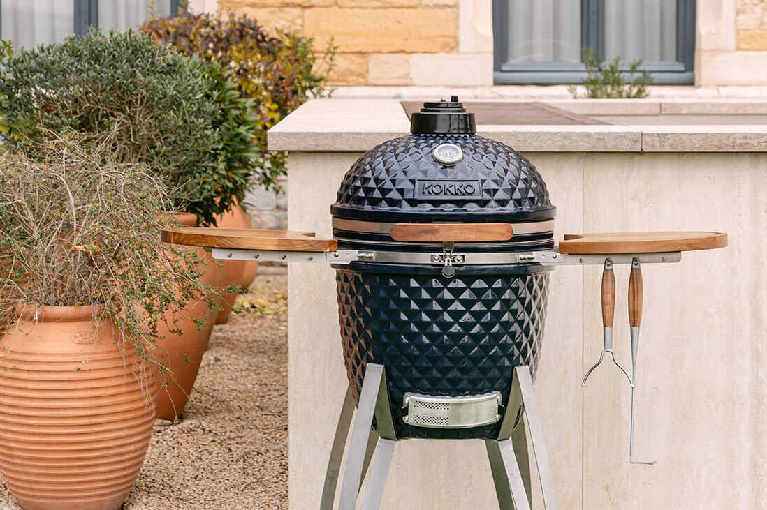 Kamado Kokko installé en extérieur, avec céramique noire texturée, tablettes latérales en bois, ustensiles suspendus et châssis métallique, intégré à un espace cuisine de jardin.