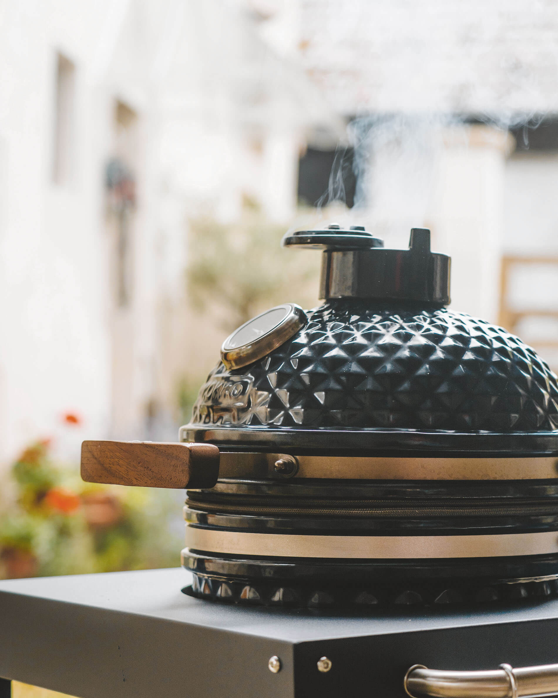 Kamado Kokko color negro sobre carro, con tapa de cerámica texturizada, asa de madera, termómetro integrado y humo saliendo de la chimenea.