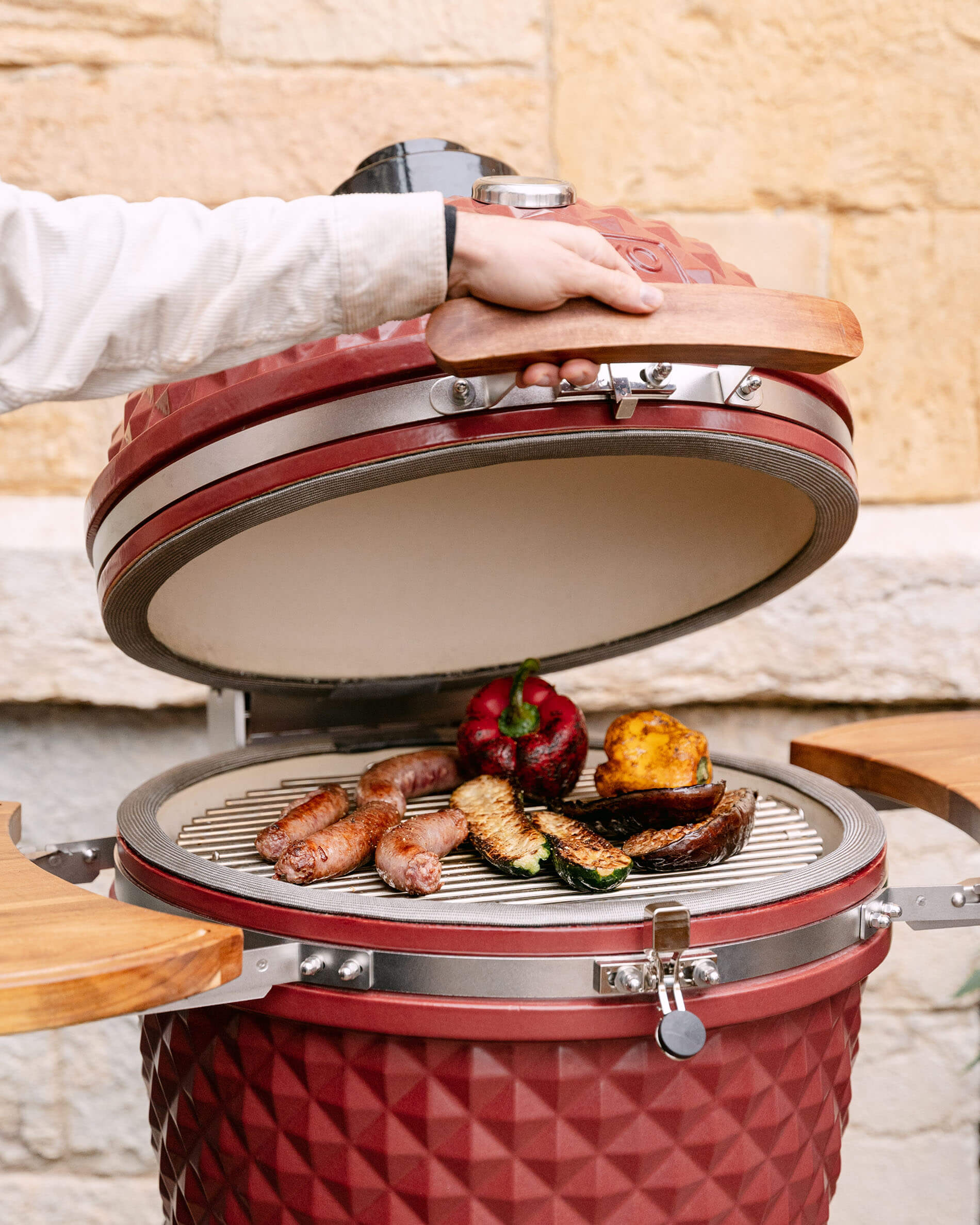Kamado Kokko de 22 pulgadas en cerámica rojo óxido abierto, cocinando salchichas y verduras a la parrilla.