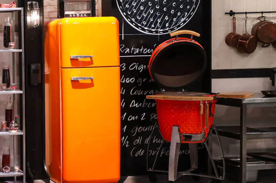 Kamado Kokko rojo de cerámica instalado en el set del programa Top Chef, con la tapa abierta y estantes laterales de madera, en una cocina profesional de rodaje.