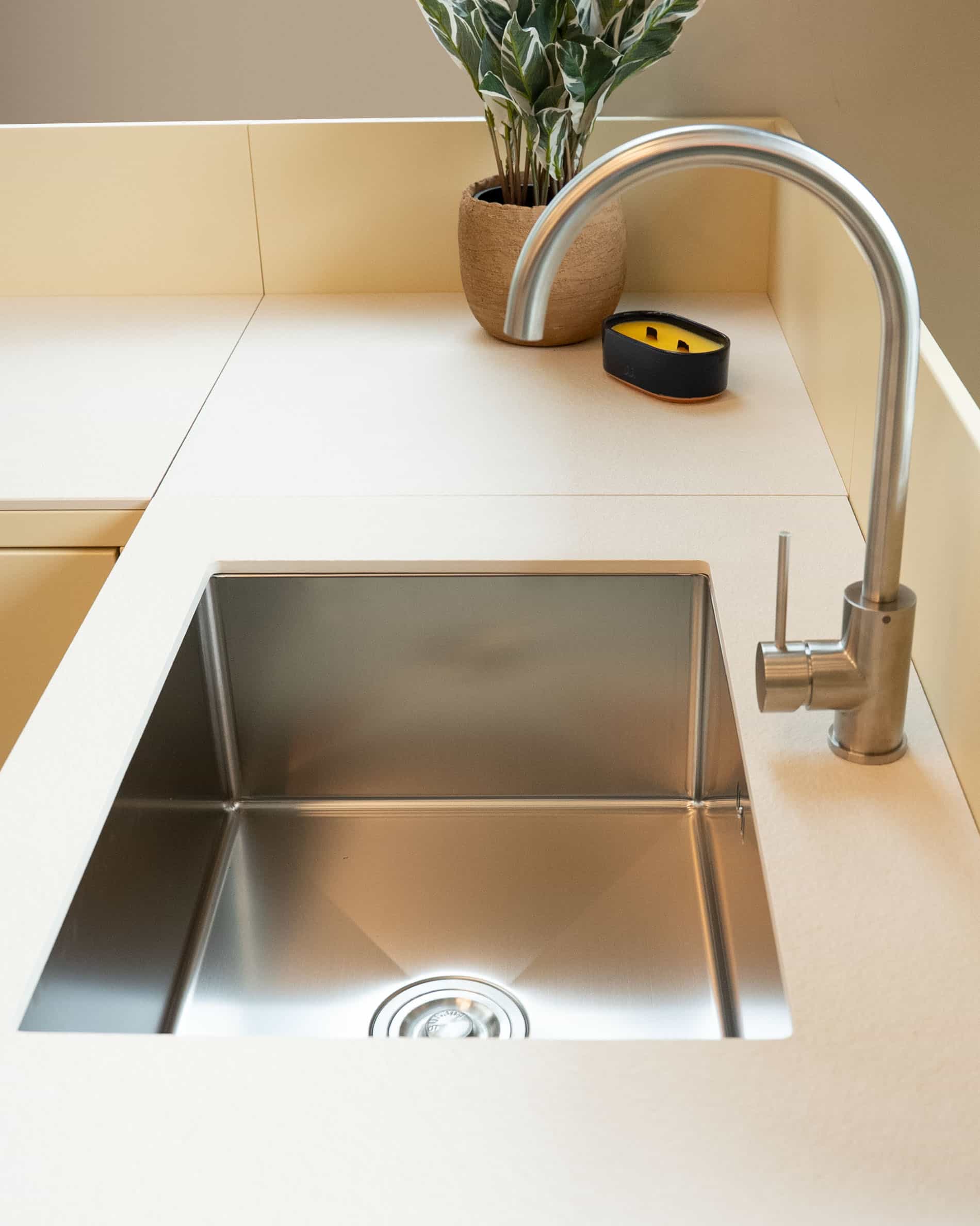 Detalle del fregadero integrado en una cocina pilotis, con cubeta de acero inoxidable, grifería metálica y encimera color crema.